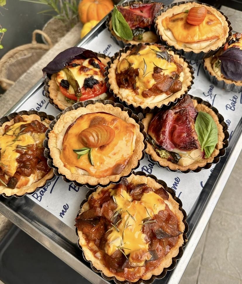 Bir tepside özenle sergilenen, üzerinde Bake Some logosu bulunan kağıtlarla sunulan, karamelize soğanlı peynirli, domatesli fesleğenli, kestane kremalı ve çeşitli mevsimlik sebzelerle hazırlanmış birçok artizan kiş ve tart, İzmir'in en iyi pastaneleri arasında yer alan Bake Some'un modern ve sürdürülebilir lezzet anlayışını yansıtıyor.
