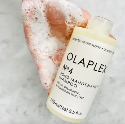Olaplex No. 4 Bond Maintenance Şampuan şişesi, beyaz köpükle kaplı bir elin yanında, mermer görünümlü bir zeminde dururken görülüyor; bu vegan saç bakım ürünü, saçları onararak daha parlak ve sağlıklı hale getirmesiyle öne çıkıyor.