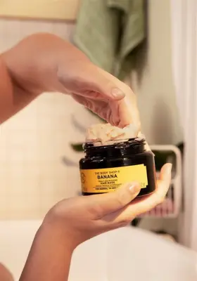 Banyo ortamında, bir kişinin eli The Body Shop Banana Saç Maskesi kavanozundan muz püresi ve Brezilya fındığı yağıyla zenginleştirilmiş krem kıvamında vegan saç bakım ürününü nazikçe çıkararak parlak ve canlı saçlara kavuşmak için kullanıma hazırlanıyor.