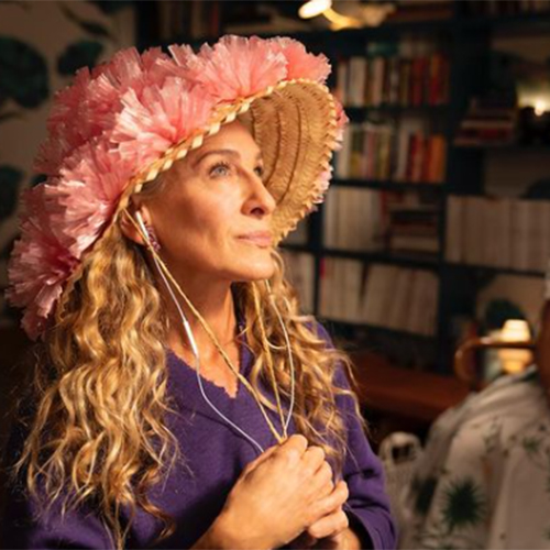 Sarah Jessica Parker'ın And Just Like That dizisindeki Carrie Bradshaw karakterini canlandırdığı bu karede, kendine özgü stilini yansıtan pembe fırfırlı süslemeli, hasır bir şapka ve beyaz kulaklıklarla, uzun kıvırcık sarı saçları dikkat çekiyor; mor V yaka kazağıyla kapalı bir ortamda kitaplık önünde dururken, dizinin ikonik kostümleri ve aksesuar tercihleri arasında yer alan bu görünüm, karakterin eklektik moda anlayışını sergiliyor.