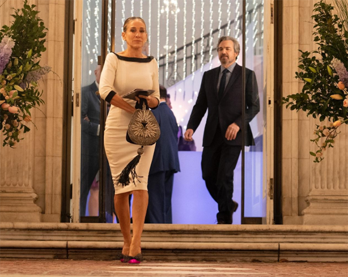 Carrie Bradshaw, And Just Like That dizisinin bir sahnesinde, siyah biye detaylı krem rengi elbisesi, işlemeli püsküllü çantası ve pembe burunlu topuklu ayakkabılarıyla şık bir binanın kapısından çıkarak dikkat çekici kostümleri sergilerken, arkasında takım elbiseli Aidan Shaw da görülüyor.