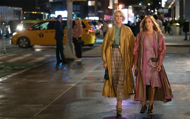 New York gecesi yağmur sonrası ıslak bir kaldırımda, And Just Like That dizisinin öne çıkan kostümleriyle özgün tarzlarını yansıtan Carrie Bradshaw, pembe ve mor tonlarındaki katmanlı elbisesiyle, yanında maskülen stile göz kırpan Miranda Hobbes ise açık kahverengi trençkotu ve desenli pileli eteğiyle yürüyor.
