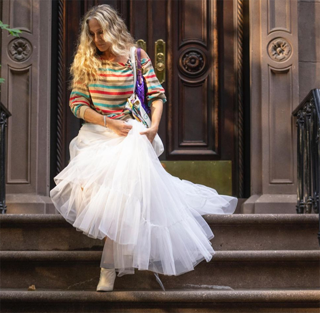 And Just Like That dizisinin dikkat çeken kostümlerinden biriyle Carrie Bradshaw, uzun dalgalı sarı saçları ve omuzları açık renkli çizgili bluzuyla, Big'i kaybettikten sonra döndüğü eski dairesinin önünde, ikonik tütü eteğe atıfta bulunan kabarık beyaz tül eteğini eliyle tutarak merdivenlerden inerken mor omuz çantası ve bej botlarıyla sade ama şık bir görünüm sergiliyor.