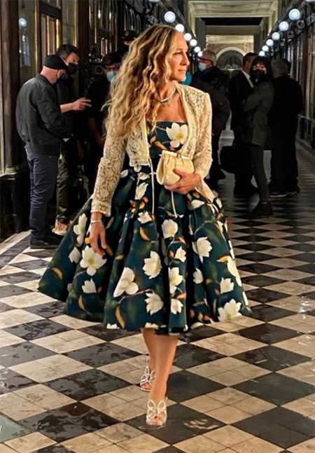 Sarah Jessica Parker, And Just Like That dizisindeki ikonik kostümlerinden biri olan, bej dantel ceketle tamamladığı, koyu yeşil üzerine büyük beyaz manolya desenli Oscar de la Renta elbisesiyle, siyah beyaz ekoseli koridorda zarifçe yürürken çekim ekibi de arkasında belirginleşiyor.