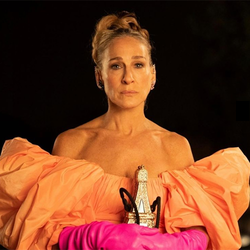 And Just Like That dizisinin final sahnesinden alınan bu yakından çekim portrede, Carrie Bradshaw karakterini canlandıran Sarah Jessica Parker, kabarık kollu şeftali tonlarında straplez elbisesi, uzun fuşya eldivenleri ve ikonik Eyfel Kulesi şeklindeki çantasıyla dikkat çeken kostümleri arasında güçlü bir duruş sergiliyor.