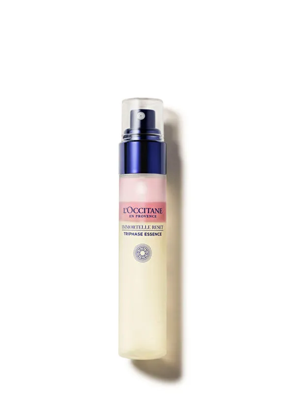 Beyaz arka plan üzerinde duran, L'Occitane Immortelle Reset Triphase Essence yazılı, sprey başlıklı, şeffaf kapaklı, koyu mavi detaylı, pembe ve krem rengi belirgin katmanları olan yenileyici ve üç fazlı bir seyahat boy essence şişesi.