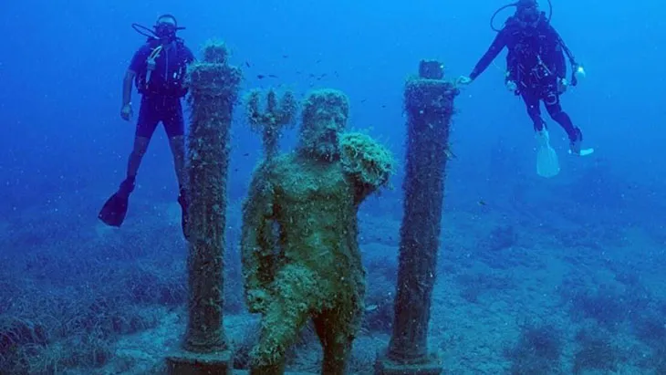 Side Su Altı Müzesi'nin derin ve berrak mavi sularında, deniz yosunlarıyla kaplanmış görkemli Deniz Tanrısı Poseidon heykeli ve onu çevreleyen antik sütunların arasında nazikçe süzülen iki dalgıç, bu eşsiz su altı dünyasının büyüleyici keşfini deneyimliyor.