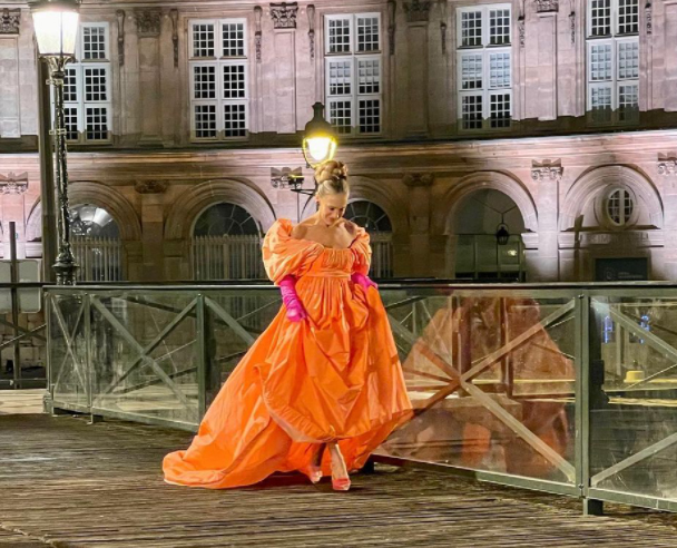And Just Like That dizisinden Carrie Bradshaw, gece ışıklarının aydınlattığı tarihi bir binanın önündeki köprüde yürürken, hacimli, omuzları açık, parlak turuncu Valentino elbisesi, uzun fuşya eldivenleri ve uyumlu topuklularıyla karakterin ikonik kostümleri arasında unutulmaz bir görünüm sergiliyor.