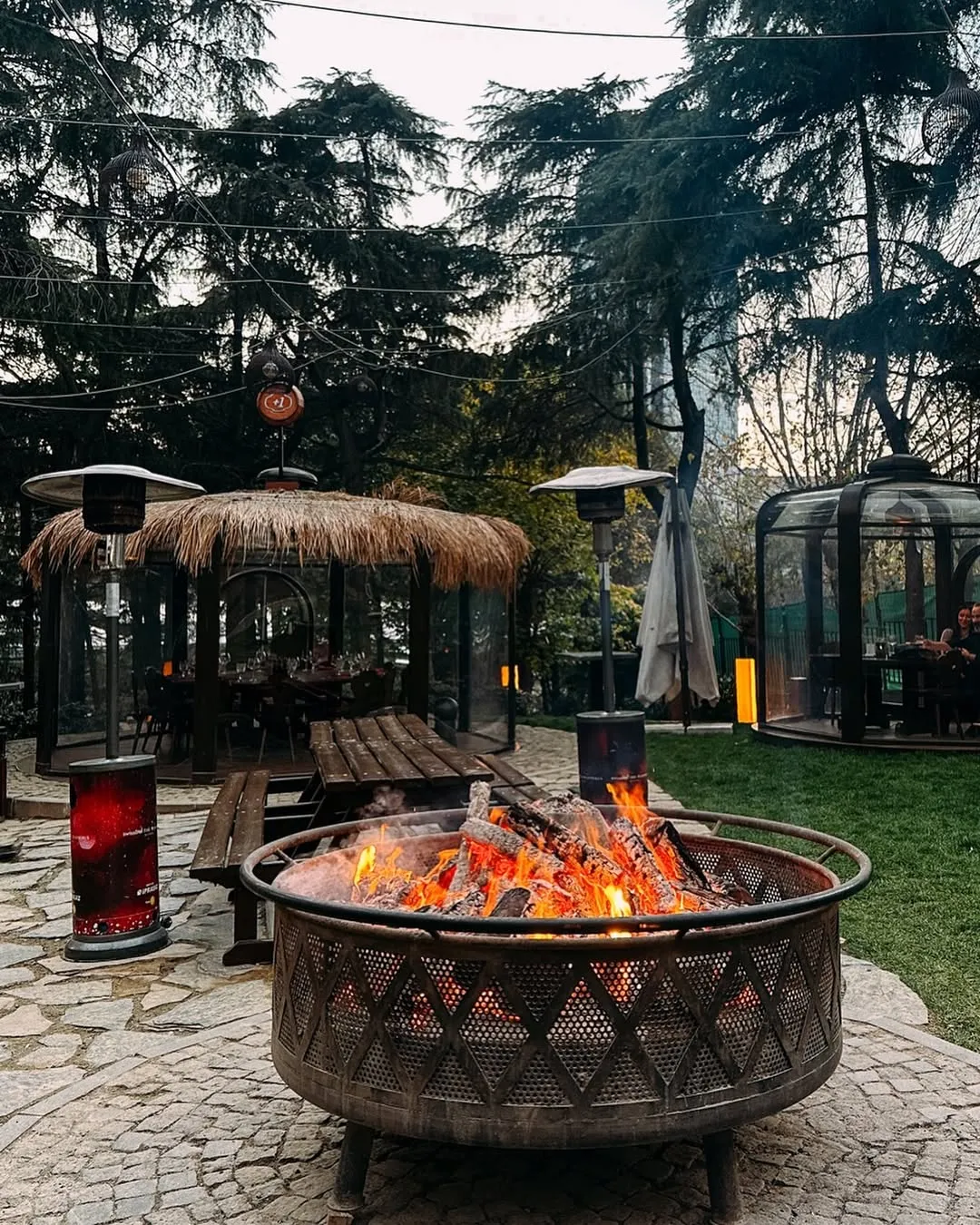 Swissôtel The Bosphorus, İstanbul'daki Chalet Garden'da yanan bir ateş çukurunun sıcak ışığında, kış atmosferini tamamlayan şık cam iglolar ve etrafındaki ağaçlar eşliğinde, Haftanın Önerileri kapsamında sunulan keyifli bir akşam yemeği ortamı.