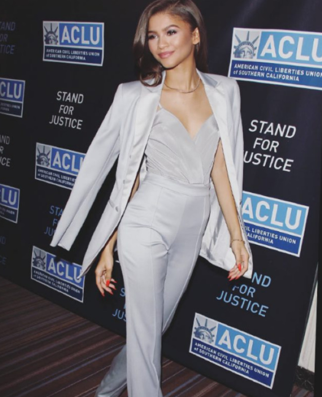 Zendaya, Beverly Hills'teki 2015 Bill of Rights yemeğinde, üzerinde ACLU logosu ve Stand For Justice yazısı olan siyah arka planın önünde, omzuna attığı ceketi, derin yakalı bluzu ve boru paça pantolonuyla pastel gümüşi tonlarda maskülen çizgiler taşıyan şık bir stil sunuyor.