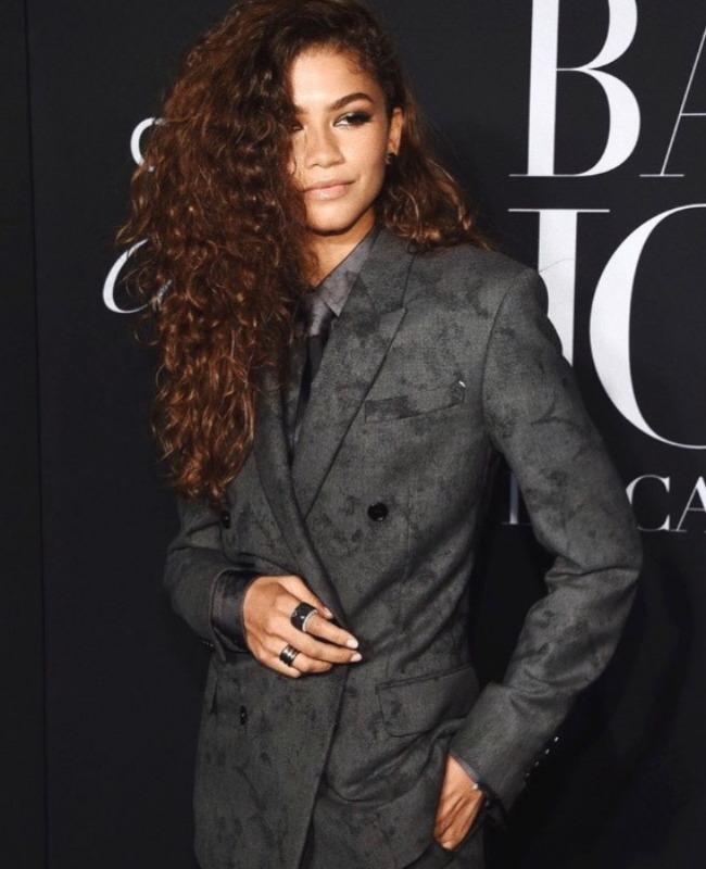 Zendaya, kıvırcık koyu kahverengi saçlarının bir kısmı yüzünü kaplarken hafifçe gülümseyerek, gri, kendinden desenli kruvaze bir takım elbiseyle kameralara poz veriyor; gri gömleğinin ve dar kravatının tamamladığı bu maskülen tavır sergileyen görünümü, geniş siyah yüzükleri ve özgün stil tercihleriyle güçlü ve cüretkar moda anlayışını yansıtıyor.