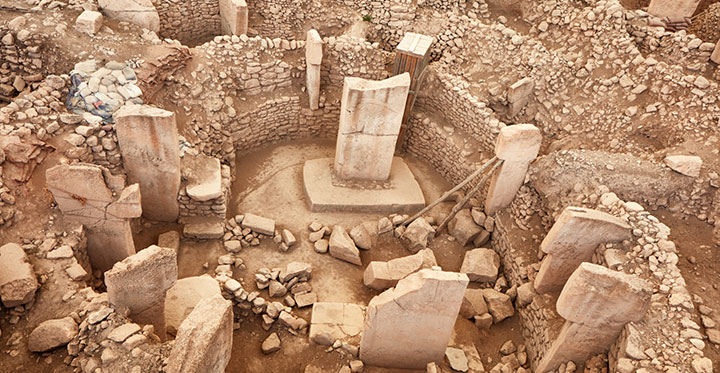 Göbeklitepe'nin kuşbakışı görüntüsünde, toprağın içinden çıkan, dairesel düzenlenmiş, yer yer sağlam, yer yer yıkılmış büyük T biçimli taş sütunlar ve çevreleyen alçak taş duvarlar, Türkiye'nin antik kentleri arasında benzersiz bir tarih öncesi yerleşim alanının kalıntılarını sergiliyor.