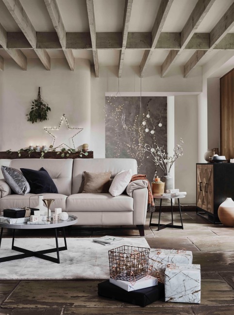 Modern bir salonda yılbaşı dekorasyonu; krem rengi kanepe, minimal çam dalları ve yıldız ışıklandırmasıyla süslenmiş duvar, mermer desenli hediye paketleri ve sıcak tonlu doğal aksesuarlarla huzurlu kış atmosferi.