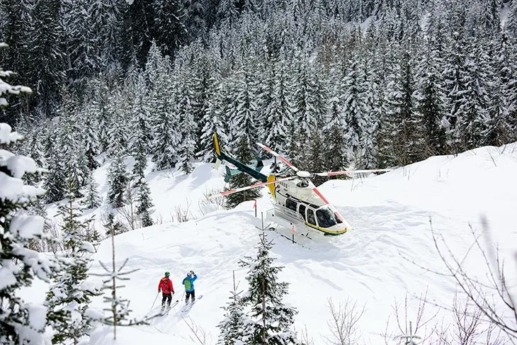 Karla kaplı dağlarda helikopterle bırakılan iki kayakçı; heliski deneyimi ve off-piste kış sporu atmosferi.