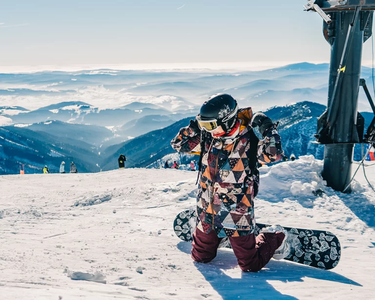 Dağ manzarası eşliğinde snowboard yapan bir kadın; renkli snowboard ceketi, kış sporu ekipmanları ve pist atmosferi.