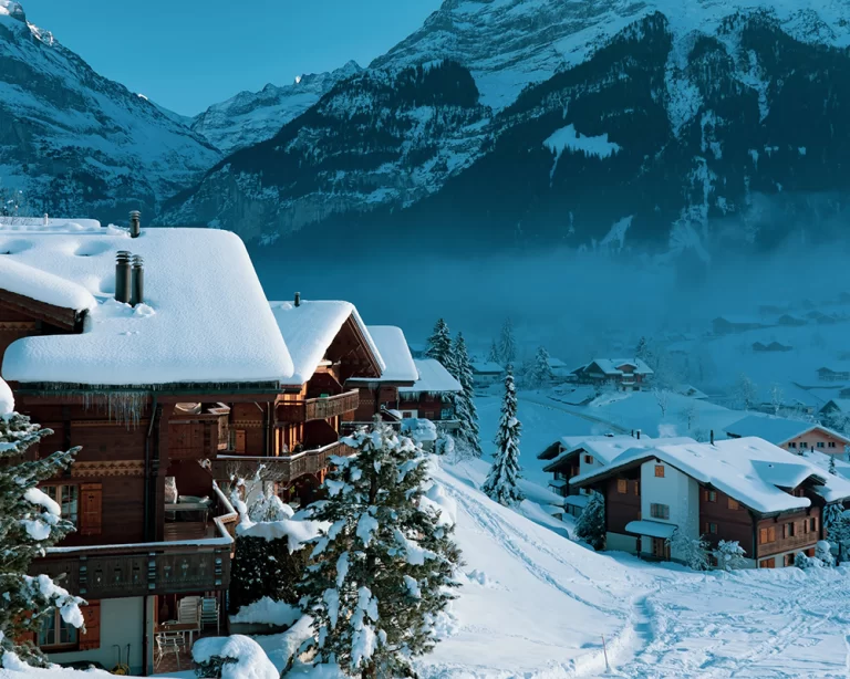 Karla kaplı Alp dağları manzarasında ahşap chalet tarzı kayak otelleri ve kış tatili köyü atmosferi.