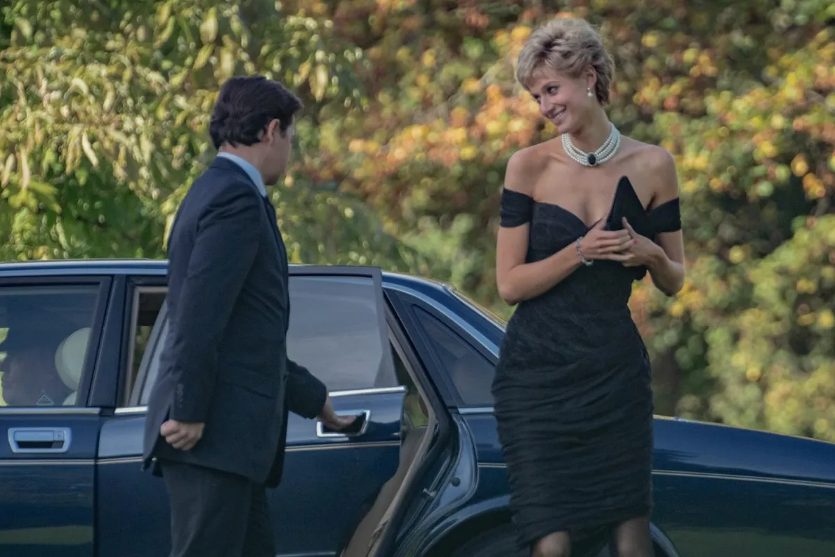 Koyu mavi bir arabanın arka kapısını açan bir adamın yanında, Elizabeth Debicki'nin canlandırdığı Prenses Diana, efsanevi omuzları açık siyah intikam elbisesi, çok katlı inci kolyesi ve elinde zarif bir portföy çantasıyla hafifçe gülümseyerek çevresine bakıyor.