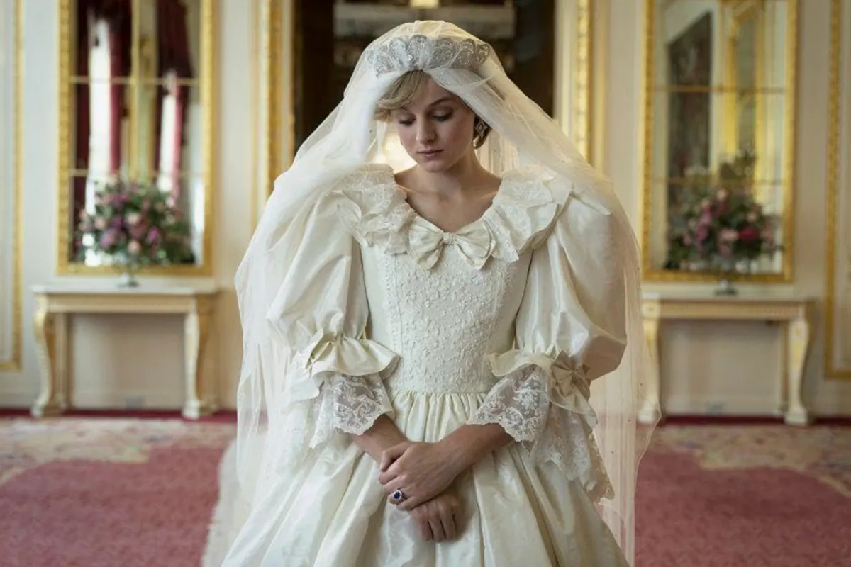 The Crown dizisinden Prenses Diana'yı canlandıran Emma Corrin, kabarık kollu, dantel detaylı ve fırfırlı yaka beyaz gelinliği, tacı ve duvağıyla başı hafif öne eğik, elleri kenetli mavi safir yüzüğüyle gösterişli bir saray odasında duruyor.
