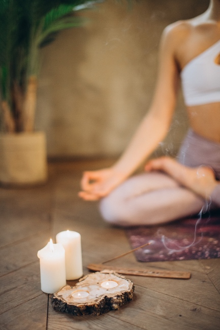 “Yere oturmuş şekilde meditasyon yapan bir kişi, önünde yanan mumlar ve tütsüyle huzurlu bir ritüel alanı oluşturmuş; zihinsel dinginlik ve farkındalık dolu bir an.”

