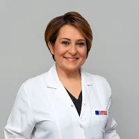Ayşe Sertkaya, beyaz önlüğü ve kısa kahverengi saçlarıyla kameraya gülümseyen bir doktor olarak görünmekte; önlüğünün yakasındaki Anadolu Sağlık Merkezi logosuyla, sade gri bir stüdyo fonunda profesyonelce poz veriyor.