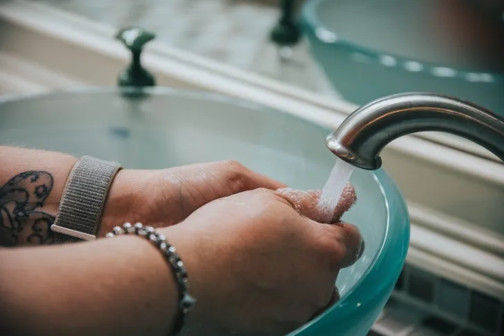 Lavabo üzerinde akan su altında ellerini yıkayan bir kişi; metal musluktan su akarken eller sabunlu şekilde ovalanıyor, arka planda seramik lavabo ve banyo detayları görülüyor.