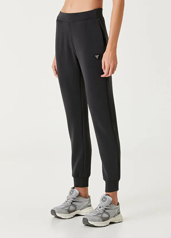 Daralan paçalı siyah bir jogger pantolon ve gri spor ayakkabılarla tamamlanmış sportif stil kombininde, pantolonun sol üst kısmında yer alan üçgen metal logo modern ve şık bir detay olarak öne çıkıyor, çabasız şıklığı vurguluyor.