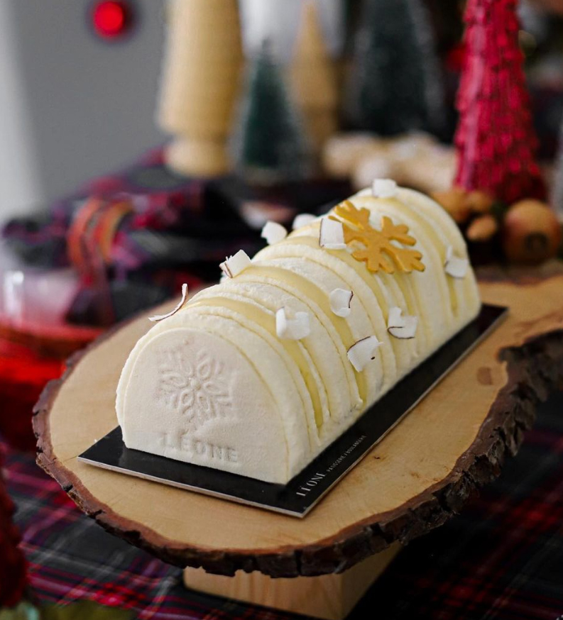 İzmir'in en iyi pastanelerinden Leone Patisserie'nin kış temalı sunumlarından, üzerinde zarif bir kar tanesi motifi ve marka adı işlenmiş, hindistan cevizi parçacıklarıyla süslü, dilimli, kütük şekilli beyaz pastası rustik bir ahşap servis tablasında sergileniyor.
