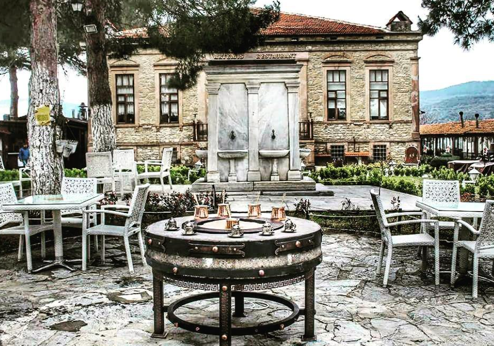 Şirince'de, tarihi Taş Mektep binasında yer alan Artemis Restoran'ın taş döşeli avlusunda, kumda Türk kahvesi için hazırlanmış bakır cezvelerle donatılmış yuvarlak bir masanın etrafındaki beyaz bahçe mobilyaları ve arka plandaki zarif mermer çeşme, bu Ege köyünde zamanın yavaşladığı huzurlu bir anı betimliyor.