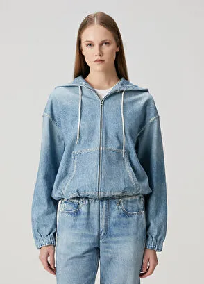 Düz açık gri bir arka planda, sportif stili vurgulayan, başlığı beyaz ipli bağcıklı, fermuarlı, beli ve bilekleri lastikli, açık mavi denim kapüşonlu bir üst ve aynı tonda bol kesim denim pantolon giymiş genç bir kadın tüm vücut poz veriyor.