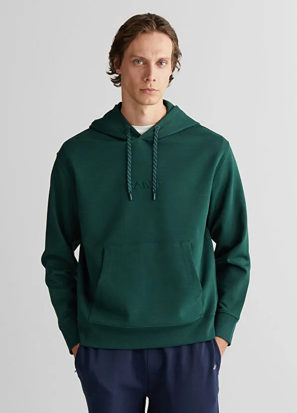 Üzerinde GANT logosu bulunan koyu yeşil kapüşonlu sweatshirt ve lacivert pantolon giyen, açık gri fon önünde duran genç bir erkek model, günlük kullanıma uygun modern ve sportif bir stil sunuyor.
