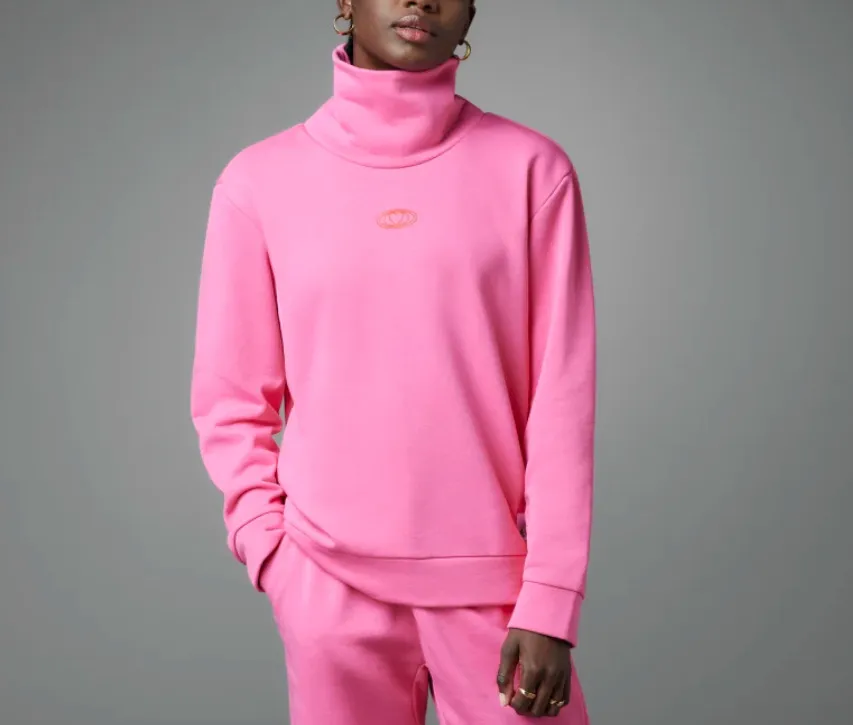 Parlak fuşya rengi, balıkçı yaka bir sweatshirt ve uyumlu eşofman altı giyen, sol elini cebine sokmuş, sade gri bir arka plan önünde duran bir kişi, rahat ve şık sportif stilini yansıtıyor.