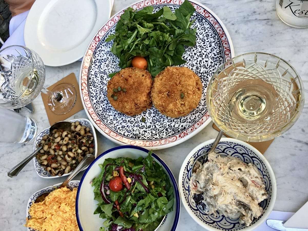 Mermer bir masanın üzerinden çekilen bu fotoğrafta, kaparili iki kızarmış köfte, taze roka salatası, börülce salatası, havuç salatası ve kremalı bir mezenin yanı sıra şarap ve su kadehleriyle donatılmış davetkar bir yemek masası, İstanbul'un bahçeli mekanlarından birinde keyifli bir buluşmayı yansıtıyor.