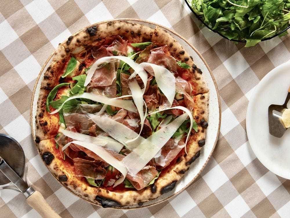 İstanbul'un en güzel bahçeli mekanlarından birinde sunulan, taze roka, füme et ve bol parmesan dilimleriyle bezenmiş, kenarları hafifçe kömürleşmiş Napoli usulü pizza, yeşillikler içindeki bir yemek deneyimi için eşsiz bir lezzet vaat ediyor.