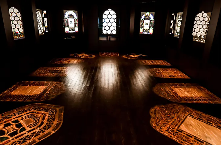 Beykoz Cam Müzesi'nin loş bir sergi salonunda, duvarlardaki karmaşık desenli vitray pencerelerden yayılan ışıkla aydınlanan ahşap zemine yansıyan geleneksel motifli halı projeksiyonları, cam sanatının büyüleyici atmosferini sergiliyor.