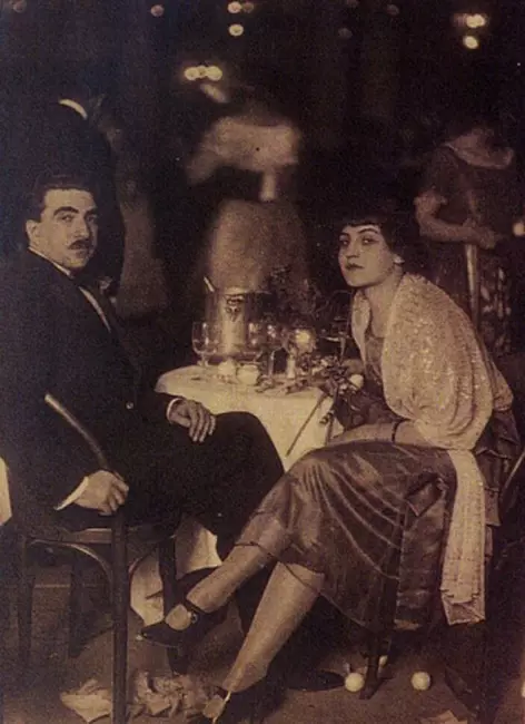 Fahrünnisa Zeyd'in sıra dışı hayatı ve sanatı bağlamında, dönemin sosyal ortamını yansıtan sepya tonlu bu fotoğrafta, koyu takım elbiseli bıyıklı bir adamla yuvarlak bir masa başında oturan, şık giyimli ve kendine özgü, doğrudan kameraya bakan genç Fahrünnisa Zeyd görünüyor, masada kadehler ve şarap kovası varken, arka plan bulanık ışıklarla dolu.