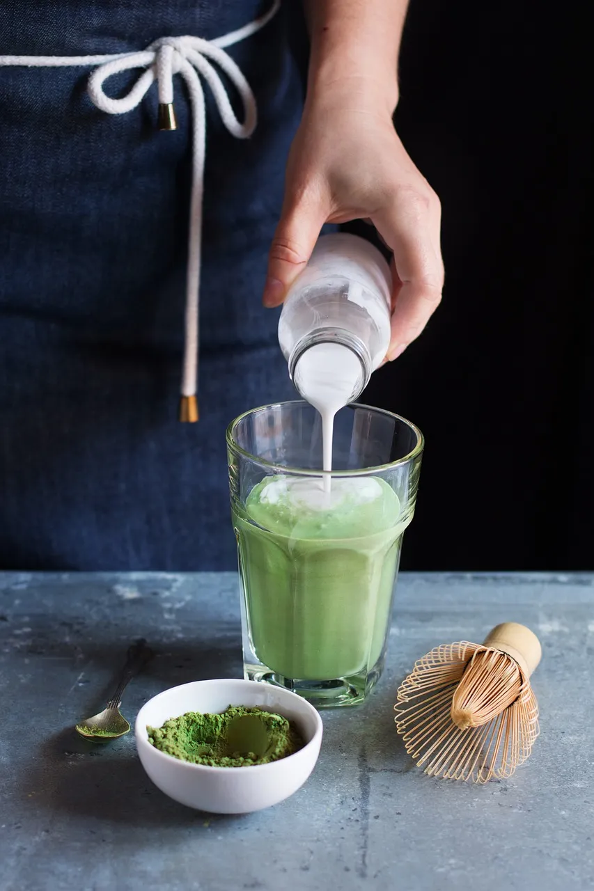 Bardakta hazırlanan matcha latteye süt eklenirken; yanında matcha tozu, küçük kaşık ve bambu çırpıcıyla sade ve modern bir hazırlık anı.