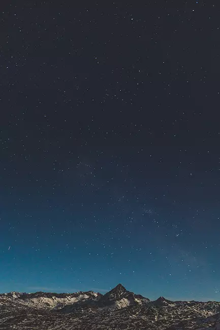 Gece karanlığında yıldızlarla kaplı gökyüzü ve karla örtülü dağ silüeti.