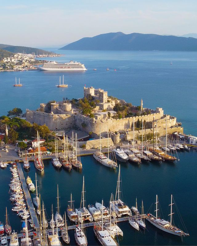 Bodrum Kalesi'nin havadan çekilmiş nefes kesici manzarasında, tarihi surları Ege'nin masmavi sularıyla buluşurken, kale çevresindeki marina lüks yatlar ve yelkenlilerle dolup taşıyor, uzakta ise bir cruise gemisi ve yarımadanın karakteristik beyaz yapıları beliriyor.
