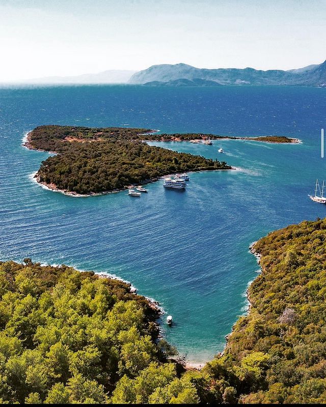 Marmaris'in berrak turkuaz sularıyla çevrili, yemyeşil ağaçlarla kaplı kıyı şeridinin ve üzerinde yüzen teknelerin havadan çekilmiş bu büyüleyici görüntüsü, bölgenin doğal güzelliğini ve tekne turlarıyla keşfedilebilecek saklı koylarını gözler önüne seriyor.