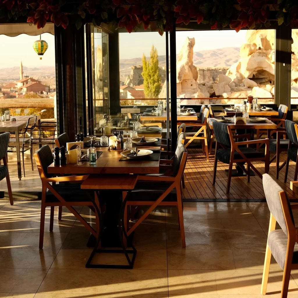 Kapadokya'da bulunan bu şık restoranın geniş pencerelerinden, masa düzenlemeleri gün batımının sıcak ışıklarıyla aydınlanırken, arka planda Çavuşin Kalesi'nin etkileyici manzarası, peribacaları ve uzakta yükselen renkli bir sıcak hava balonuyla birlikte unutulmaz bir akşam yemeği deneyimine davet eden atmosferi yansıtılıyor.
