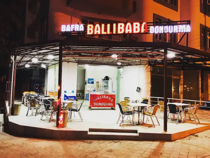 Gece ışıkları altında parlayan Bafra Ballı Baba Dondurma tabelasının önünde, açık havada metal çatılı terasında boş masa ve sandalyelerin düzenli bir şekilde sıralandığı, Samsun'un en beğenilen mekanlarından biri olan samimi bir dondurmacı görünümü.