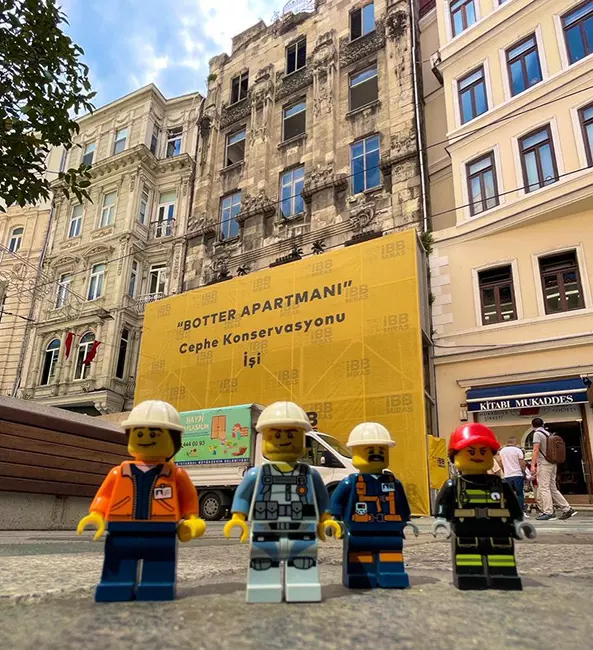 Botter Apartmanı'nın cephe konservasyonu işini duyuran büyük sarı bir pankartın önünde, çeşitli iş kıyafetleri giymiş dört Lego figürü dikkat çekiyor; bu detay, tarihi yapının restorasyon sürecine dair merak edilen bilgileri ve önemini vurguluyor.