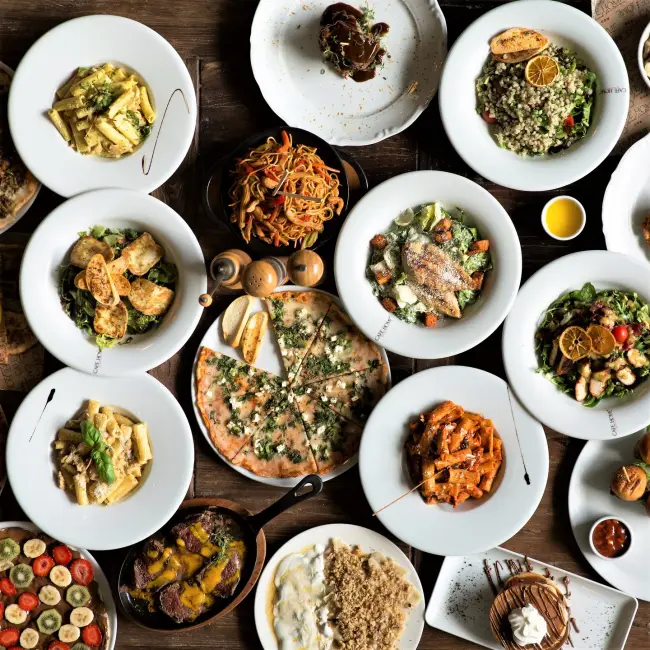 Ahşap bir masa üzerinde, Samsun'un en beğenilen mekanlarından Cafe Home'da sunulan salata, makarna, pizza, et yemeği, noodle ve tatlılar gibi günün her saatine uygun çok çeşitli lezzetlerin beyaz tabaklarda iştah açıcı bir şekilde servis edildiği, dolu dolu bir sofra manzarası.