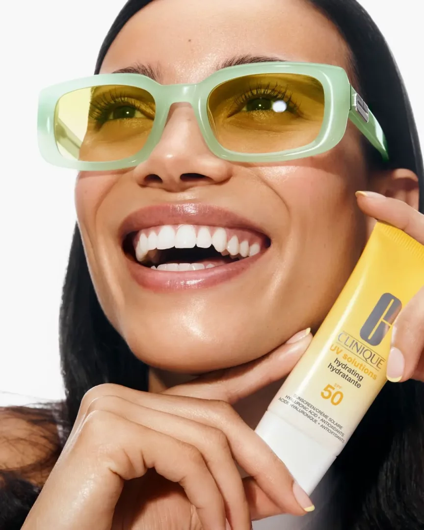 Açık yeşil renkli, sarı camlı güneş gözlüğü takmış, parlak beyaz dişleriyle içten gülümseyen bir kadın, elinde Clinique UV Solutions Hydrating Hydratante SPF 50 güneş kremi tüpünü yüzüne yakın bir şekilde gösteriyor.