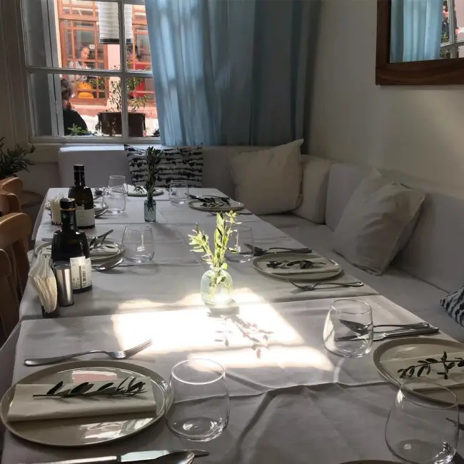 Ayvalık'ın tarihi dokusunu yansıtan bir restoranda, güneş ışıklarıyla aydınlanan beyaz örtülü uzun bir yemek masası, her tabağın üzerindeki taze zeytin dalları ve Kuzey Ege'nin zeytinyağı kültürünü çağrıştıran şişelerle sade ve davetkar bir ziyafet ortamı sunuyor.