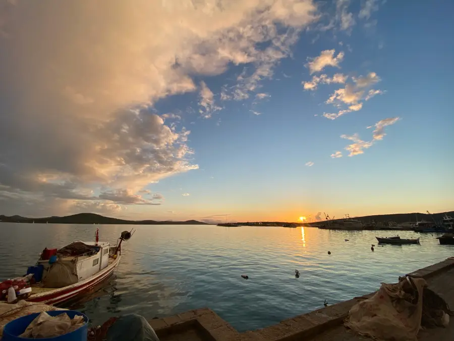 Batan güneşin turuncu tonlarıyla parlayan Kuzey Ege sularında, Ayvalık'ın Cunda sahilinde demirli bir balıkçı teknesi ve uzaktaki adalar, gökyüzündeki dramatik pembe ve turuncu bulutlarla çevrili huzurlu bir gün batımı anını yansıtıyor.