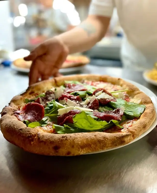 Ayvalık'ın zengin mutfağını farklı lezzetlerle taçlandıran Cunda Uno Pizza'dan, fırından yeni çıkmış, kenarları çıtır, üzerinde bolca taze roka, ince dilimlenmiş füme et ve rendelenmiş peynir bulunan iştah açıcı bir pizza, mutfak tezgahında servis edilmeye hazır bekliyor.