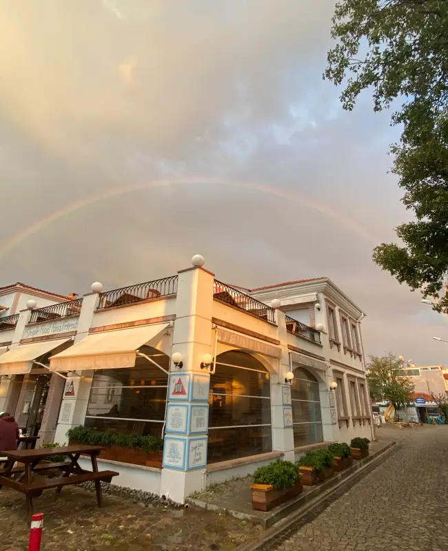 Cunda Uno Pizza restoranının önünde, ahşap piknik masalarının yer aldığı taş döşeli bir alanda, gün batımı ışıklarıyla aydınlanmış beyaz binanın üzerinde tam bir gökkuşağı beliriyor; bu Kuzey Ege güzeli Ayvalık'ta unutulmaz anlar vadeden keyifli bir sahne.