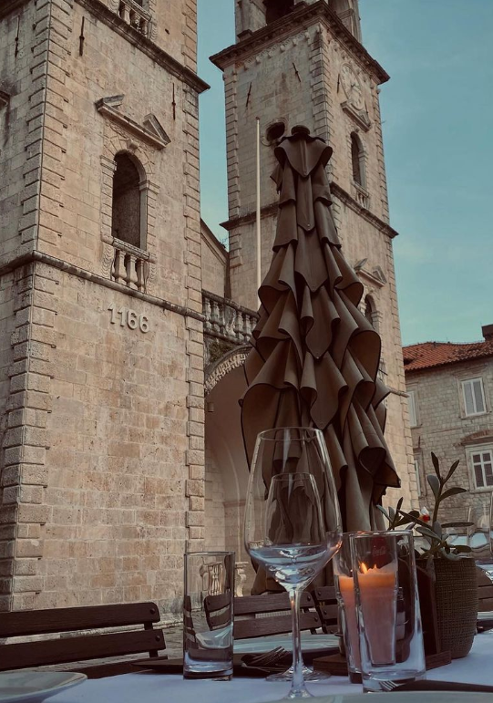 Karadağ'ın tarihi Kotor Eski Şehir'de, taş binaların ve iki görkemli çan kulesinin gölgesinde kurulmuş bir restoranda, kadehler ve mumlarla hazırlanmış bir masanın yakın çekimi, 1166 tarihli duvar detayıyla zenginleşen bu Adriyatik kasabasının eşsiz atmosferini yansıtıyor.