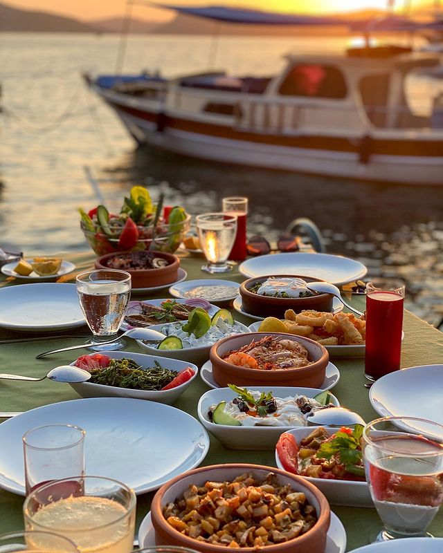 Gün batımının kızıl tonlarında, Marmaris'in sakin bir koyunda denize sıfır bir masada sunulan taze Ege mezeleri, kalamar ve karides güveç gibi deniz ürünleriyle dolu zengin bir yemek ziyafeti, unutulmaz bir Marmaris tatili deneyimini vurguluyor.